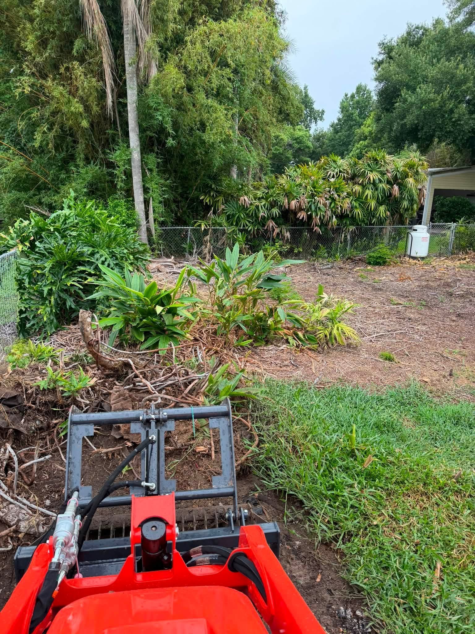 Compact loader attachment gathering roots and brush beside a fence.