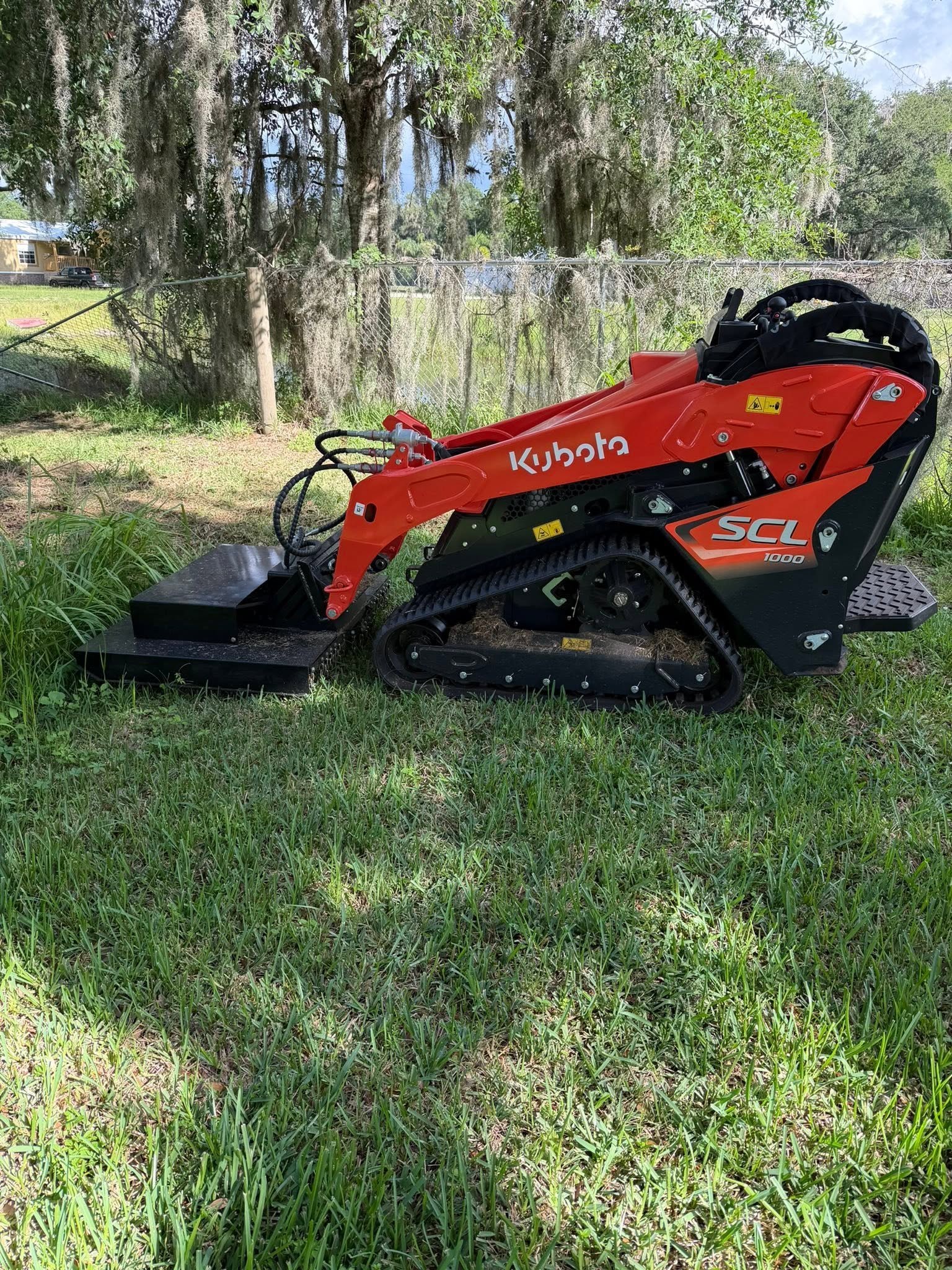 Orange compact track loader with a mower attachment in a grassy yard.