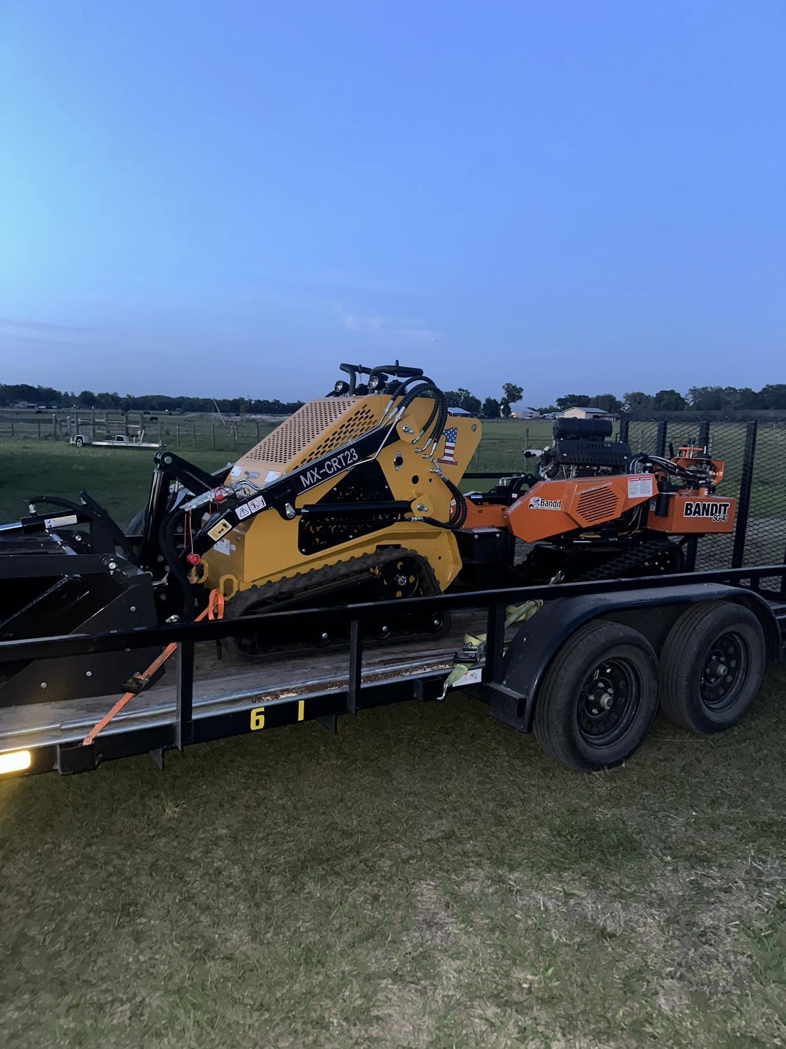Compact loader and stump grinder secured on an equipment trailer at dusk.